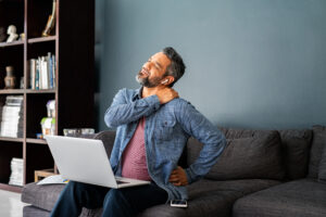 Mature indian man suffering from shoulder and back pain while sitting on couch and working from home on laptop. Indian middle aged business man in casual clothing stretching neck while working on laptop. Stressed middle eastern businessman suffering from neck pain.