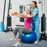 Physiotherapist working with mature female patient in a rehab center. Chinese woman lifts dumbbells on a fitness ball with a personal trainer