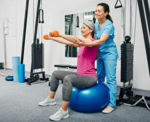 Physiotherapist working with mature female patient in a rehab center. Chinese woman lifts dumbbells on a fitness ball with a personal trainer